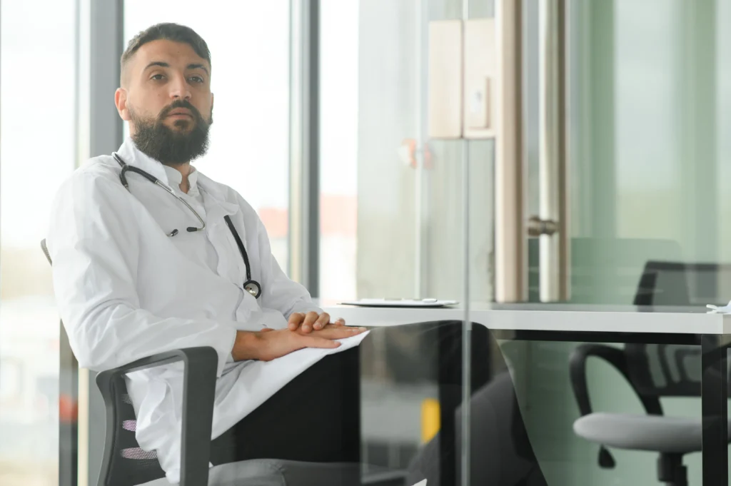 Médico com jaleco branco, homem, barbudo, sentado a cadeira de seu escritório