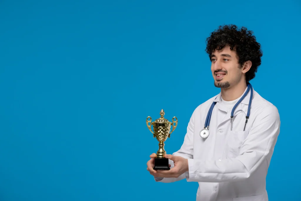 Médico branco, com cabelos encaracolados e barba pretas, com jaleco branco e segurando um troféu com as duas mãos