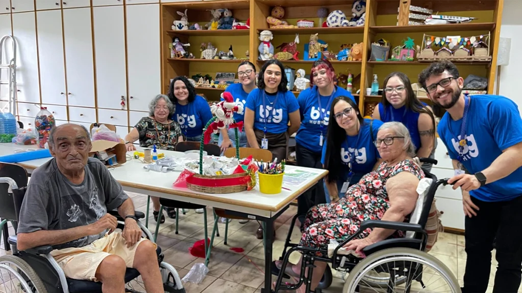 Time de pessoas com uniforme azul da BNG Hub com algumas pessoas da terceira idade, sendo um homem na esquerda, sentado em uma cadeira de rodas e uma mulher na direita, também sentada em uma cadeira.